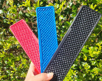 Three textured rubber mats in red, blue, and black held by a hand with a green leafy background.